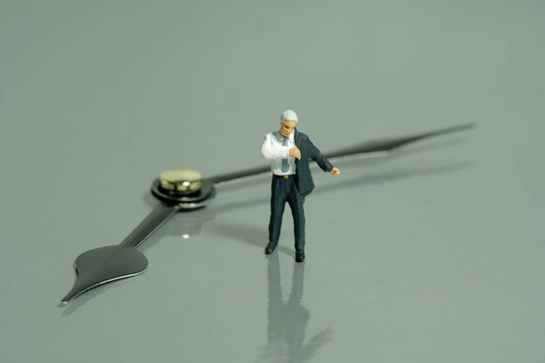 Miniature tiny people toy figure photography. Go to work concept. A businessman getting ready wearing suit in front of clockwise. Isolated on grey background. Image photo - Photo, Image