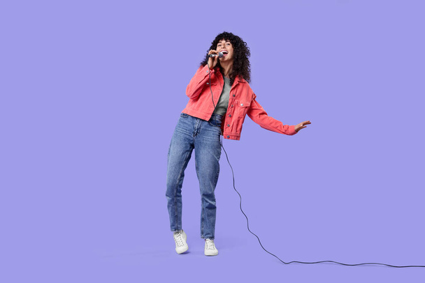 Beautiful young woman with microphone singing on purple background - Φωτογραφία, εικόνα
