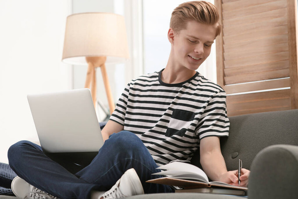 Online learning. Smiling teenage boy with laptop writing in notebook on sofa at home - Photo, Image