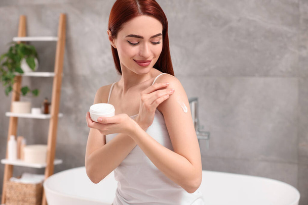 Beautiful young woman applying body cream onto shoulder in bathroom - Foto, Imagen