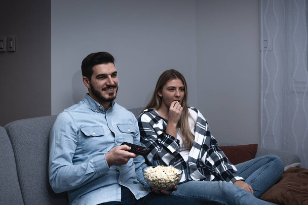 Watching TV at home. Loving couple resting on sofa and eating popcorn while enjoying movie or film on television - Fotografie, Obrázek