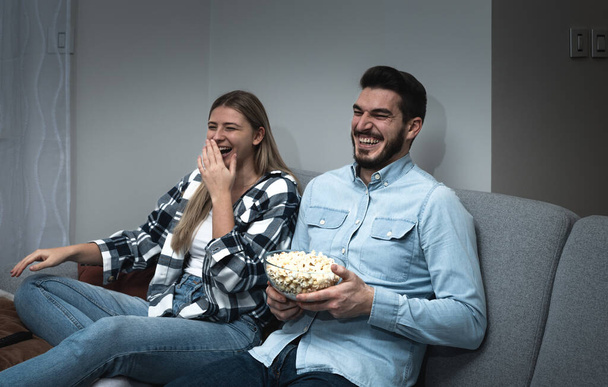 Watching TV at home. Loving couple resting on sofa and eating popcorn while enjoying movie or film on television - Fotografie, Obrázek