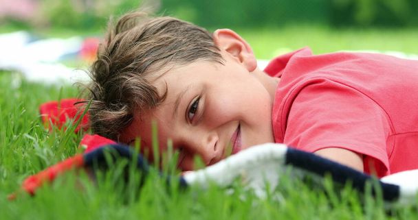 Bambino sdraiato su erba all'aperto guardando alla fotocamera. Giovane ragazzo che riposa fuori - Foto, immagini