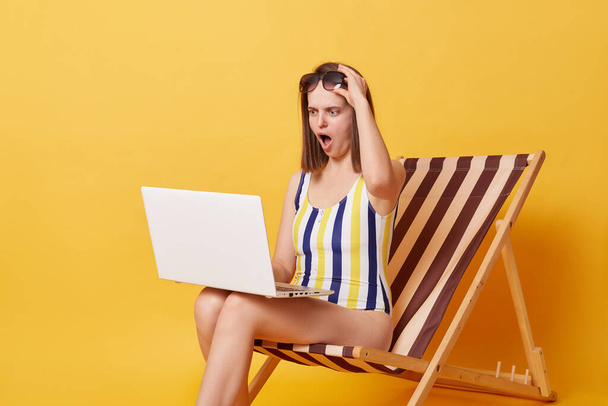 Shocked surprised young woman wearing swimsuit sitting on chair working on laptop isolated over yellow background sees astonished information browsing internet. - Valokuva, kuva
