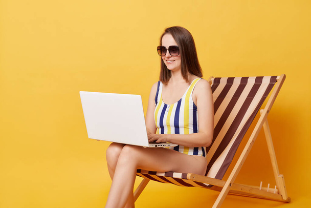 Happy young woman wearing striped one-piece swimsuit sitting on chair working on laptop pc isolated over yellow background, doing her online work. - Foto, Imagen