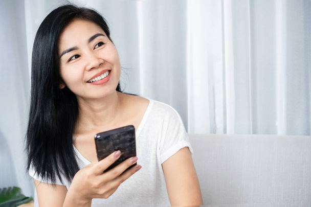 sonriente mujer asiática usando un smartphone, pensando y soñando despierto con algo con cara feliz - Foto, imagen