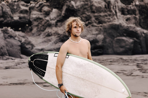 Fit jeune surfeur homme aux cheveux blonds bouclés avec planche de surf passe par l'océan en s'amusant à faire des sports nautiques extrêmes, surf. Voyage et mode de vie sain concept. Destination de voyage sportif - Photo, image