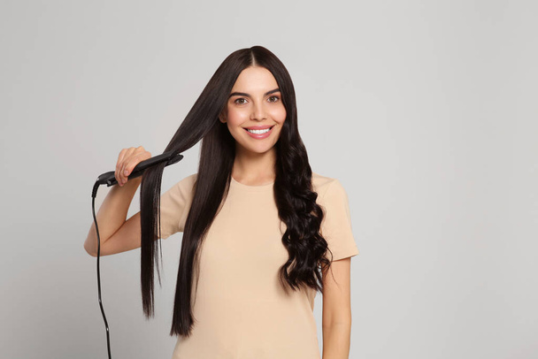 Beautiful happy woman using hair iron on light grey background. Space for text - Foto, afbeelding