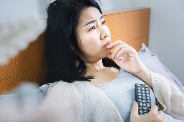 emotional Asian woman watching sad movie on TV hand holding remote control in bed - Photo, Image