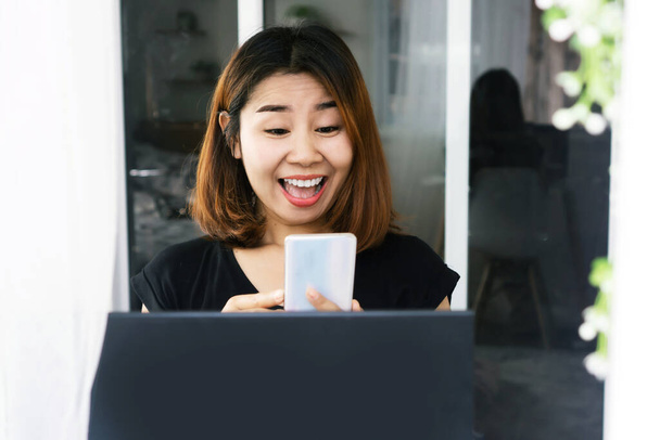 emocionado mujer asiática mano celebración de teléfono móvil con la cara sonriente obtener buenas noticias sentado con el ordenador portátil en el escritorio - Foto, imagen