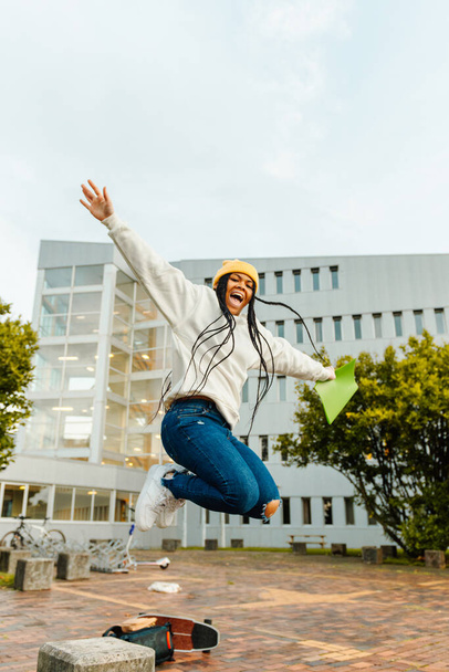 Młoda czarna dziewczyna z warkoczami skacze świętując zakończenie egzaminów przed wakacjami u bram szkoły średniej lub kampusu uniwersyteckiego. Koniec klas - Zdjęcie, obraz