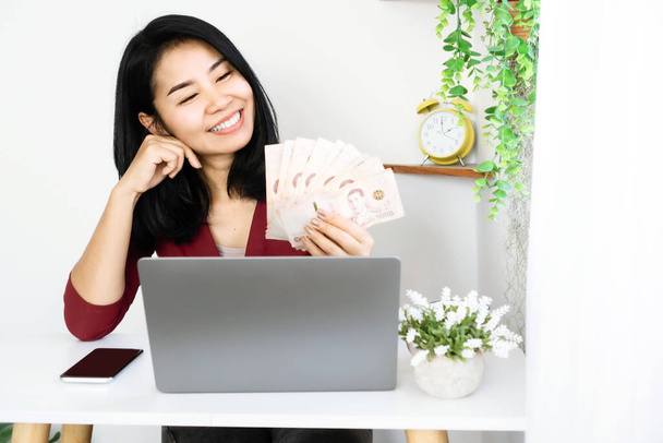 Mujer de negocios asiática sosteniendo billetes tailandeses, exitosa con hacer dinero en línea sentado con computadora portátil y teléfono móvil en el escritorio - Foto, imagen