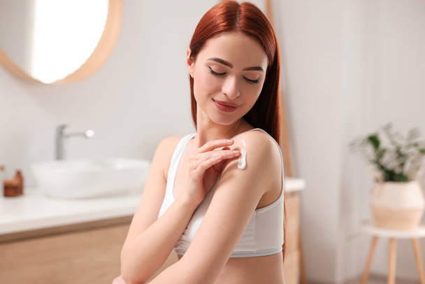 Beautiful young woman applying body cream onto shoulder in bathroom - Foto, afbeelding