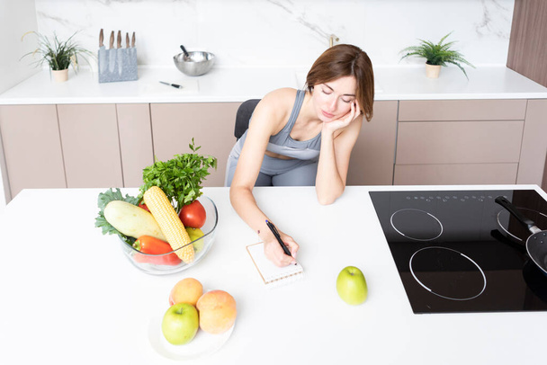 Молодая привлекательная и стройная женщина сидит за столом на кухне и пишет свой план диеты в блокноте. Много фруктов и овощей, которые на столе также включены в ее рацион - Фото, изображение