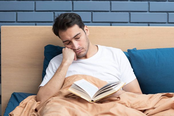 Ο κουρασμένος άντρας αποκοιμήθηκε ενώ διάβαζε το βιβλίο στο κρεβάτι. - Φωτογραφία, εικόνα