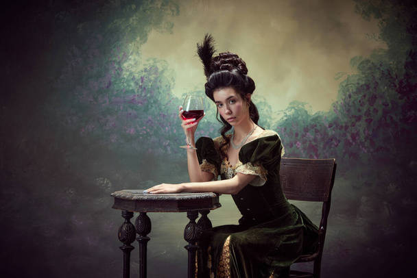 Portrait of attractive young woman, princess, queen in retro dress sitting at table with glass of red wine against dark vintage background. Concept of history, renaissance art, comparison of eras - Photo, Image
