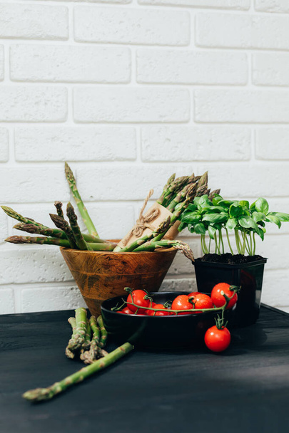 Fondo alimentario saludable. Verduras frescas, espárragos en plato de madera, tomates en plato negro y basílica verde sobre fondo de madera blanca. Puesta plana - Foto, imagen