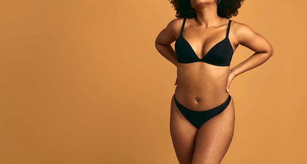 Crop African female wearing black bra and panties standing against orange background concept of self acceptance and body positive - Photo, Image