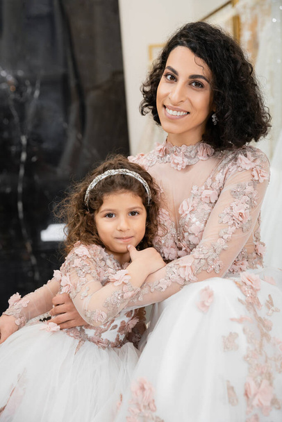 brunette middle eastern woman with wavy hair hugging cute girl and smiling near white wedding dresses in bridal salon, floral, mother and daughter, happiness, shopping, wedding day  - Photo, image