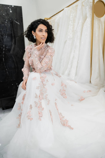 happy middle eastern woman with wavy hair sitting in gorgeous and floral wedding dress near blurred and white gown inside of luxurious bridal salon, charming and elegant, bride-to-be - Foto, imagen