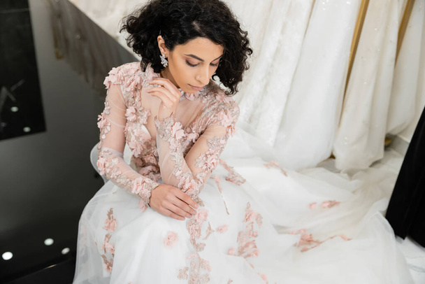 overhead view of brunette middle eastern woman with wavy hair sitting in gorgeous and floral wedding dress near blurred and white gown inside of luxurious bridal salon, shopping, bride-to-be - Fotografie, Obrázek