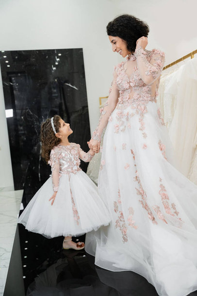cheerful middle eastern woman with wavy hair in stunning wedding dress holding hands with daughter in cute floral attire while standing in bridal salon, shopping, luxurious, golden accents  - Foto, immagini