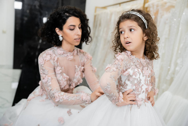 blurred middle eastern bride with brunette wavy hair in wedding dress adjusting cute floral dress of surprised daughter in bridal salon, shopping, special moment, hands on hips, white gown - Photo, Image
