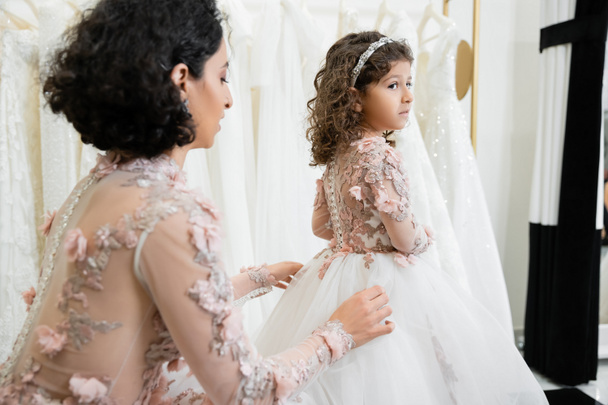 middle eastern bride with brunette wavy hair in floral wedding dress adjusting tulle skirt of daughter in cute attire in bridal salon, shopping, special moment, togetherness, white gown, festive - Foto, afbeelding
