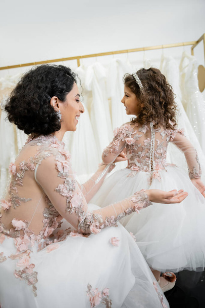 happy middle eastern woman with brunette wavy hair in floral wedding dress looking at tulle skirt of smiling daughter in cute attire in bridal salon, shopping, special moment, togetherness  - Fotografie, Obrázek