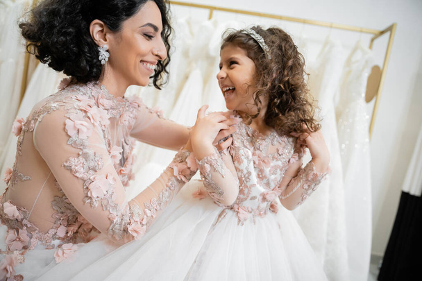 happy middle eastern woman with brunette hair in floral wedding dress hugging shoulders of smiling daughter in cute attire with tulle skirt in bridal salon, shopping, special moment, togetherness  - Foto, imagen