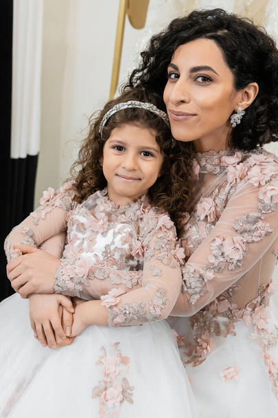 cheerful middle eastern bride in wedding dress hugging happy girl in cute floral attire in bridal salon, shopping, special moment, mother and daughter, happiness, looking at camera - Photo, Image