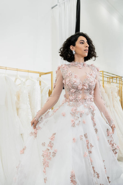 enchanting and middle eastern woman trying on gorgeous and floral wedding dress inside of luxurious bridal salon, shopping, bride-to-be,  blurred white gown on background, looking away  - Photo, Image