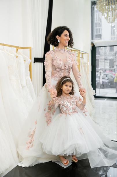 χαριτωμένο κορίτσι της Μέσης Ανατολής σε floral ενδυμασία κρατώντας τα χέρια με χαρούμενη γυναίκα στέκεται στο νυφικό κοντά θολή λευκό φόρεμα στο εσωτερικό του νυφικού κομμωτηρίου, ψώνια, νύφη-να-να, μητέρα και κόρη  - Φωτογραφία, εικόνα