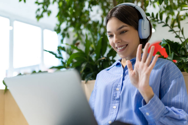Waist-up portrait of a cheerful woman in the headphones waving at someone while looking at the computer monitor - 写真・画像