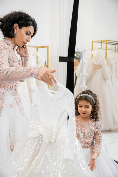 брюнетка середня східна наречена у квітковому весільному сукні допомагає вибрати сукню для своєї маленької дочки в весільному салоні навколо білих тканин тюлю, процес приготування, безтурботність
  - Фото, зображення
