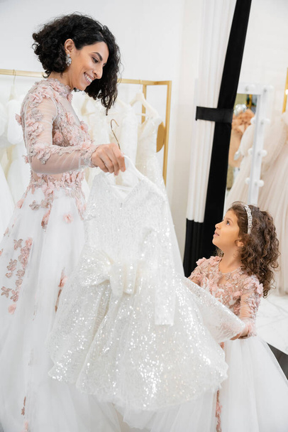 happy and brunette middle eastern bride in floral wedding gown helping to choose dress for her little daughter in bridal salon around white tulle fabrics, process of preparation, togetherness  - Photo, Image