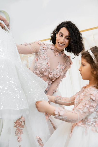 charming and brunette middle eastern woman in floral wedding gown helping to choose dress for her little daughter in bridal salon around white tulle fabrics, process of preparation, togetherness  - Photo, Image