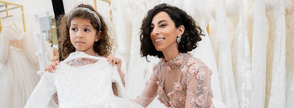 czarujący i brunetka Bliski Wschód panna młoda w kwiatowej sukni ślubnej pomaga wybrać sukienkę dla swojej córeczki w salonie ślubnym wokół białych tiulu tkanin, proces przygotowania, sztandar  - Zdjęcie, obraz