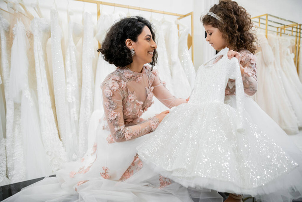 błogi i brunetka bliskowschodnia panna młoda w kwiatowej sukni ślubnej pomaga wybrać sukienkę dla swojej uroczej córeczki w salonie ślubnym wokół białych tiulu tkanin, proces przygotowania  - Zdjęcie, obraz