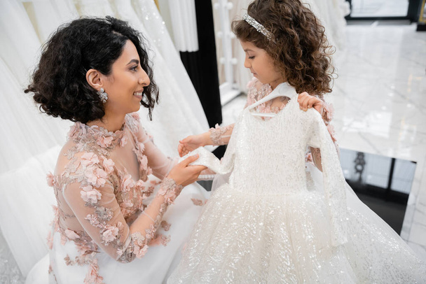 幸せとブルネット中東の花嫁で花のガウンを着て、彼女の小さな娘が白いチュール生地の周りのブライダルサロンでドレスを選択するのを助ける、結婚式の準備のプロセス  - 写真・画像