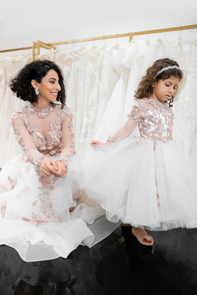 különleges pillanat, vidám közel-keleti menyasszony virágos esküvői ruha ül, és nézi a kislány menyasszonyi szalonban pózol körül fehér tüll szövet, menyasszonyi vásárlás, összetartás  - Fotó, kép