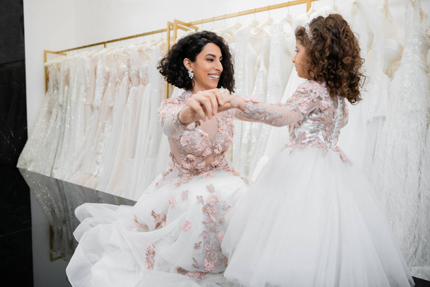 különleges pillanat, boldog közel-keleti menyasszony virágos esküvői ruhában ül és fogja a kezét a kislányával menyasszonyi szalonban a fehér tüll szövet körül, menyasszonyi vásárlás, összetartás  - Fotó, kép