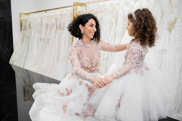 különleges pillanat, boldog közel-keleti menyasszony esküvői ruhában ül és kézen fogva kislányával menyasszonyi szalonban, fehér tüll szövet körül, menyasszonyi vásárlás, összetartás  - Fotó, kép