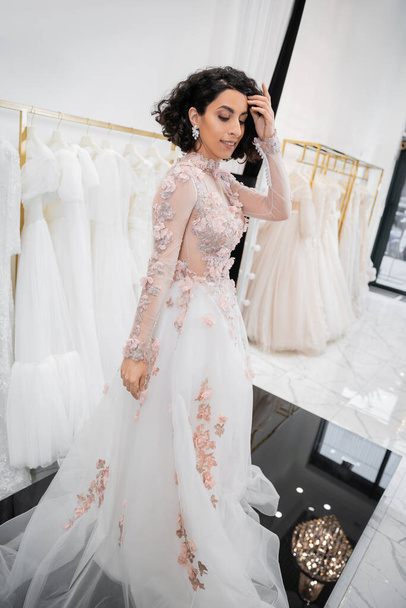 bride-to-be, charming middle eastern and brunette woman with wavy hair standing in gorgeous and floral wedding dress inside of luxurious bridal salon around white tulle fabrics, shopping  - Photo, Image