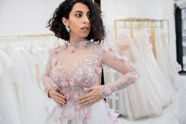 stunning middle eastern and brunette bride with wavy hair standing in gorgeous and floral attire inside of luxurious bridal salon around white tulle fabrics, wedding dress shopping  - Foto, Imagen