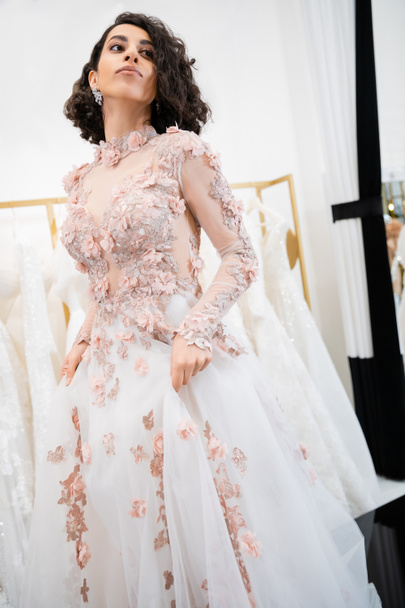 attractive middle eastern woman with wavy hair standing in gorgeous and floral wedding dress and looking away inside of luxurious salon around white tulle fabrics, bridal shopping, bride-to-be - 写真・画像
