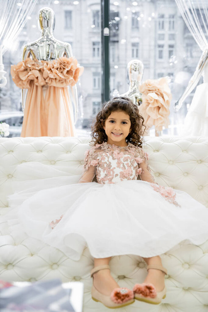 alegre chica de Oriente Medio con el pelo rizado sentado en vestido floral en el sofá blanco dentro del lujoso salón de bodas, niño sonriente, falda de tul, nupcial, maniquí borroso en el fondo  - Foto, Imagen