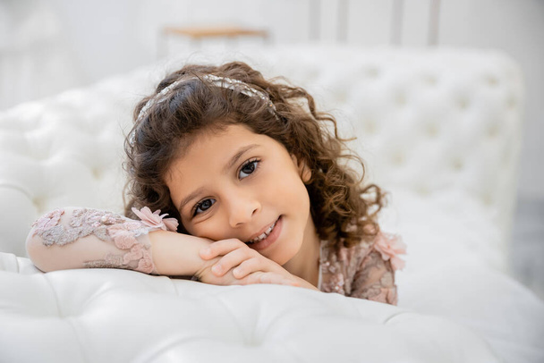 portrait of positive middle eastern girl with brunette curly hair posing in floral dress and leaning on white couch inside of luxurious wedding salon, smiling kid, blurred background  - Photo, Image