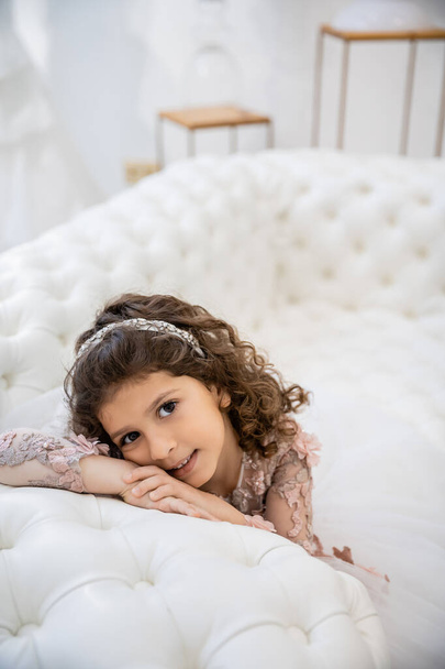 portrait of cheerful middle eastern girl with brunette curly hair posing in floral dress and leaning on white couch inside of luxurious wedding salon, smiling kid, blurred background, joy  - Photo, Image
