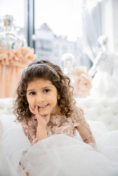 cheerful middle eastern girl with curly hair posing in floral dress with tulle skirt and sitting on white couch inside of luxurious wedding salon, smiling kid, blurred background  - Photo, Image
